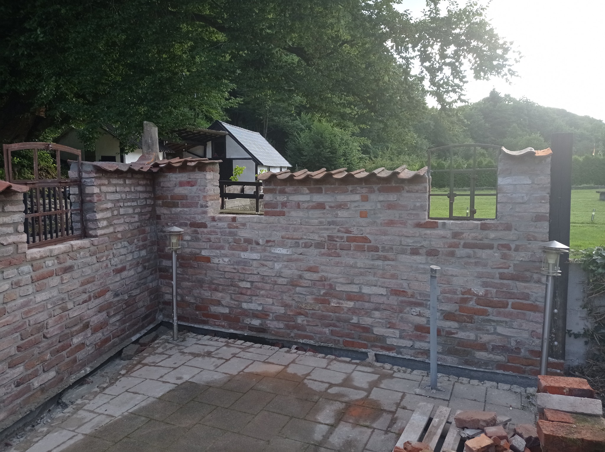Murowana z czerwonej cegły ściana z fragmentami metalowych elementów i ceramicznymi dachówkami na górze, widoczne lampy ogrodowe i pozostałości materiałów budowlanych na placu.