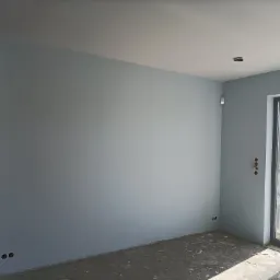 Pomalowane na jasnoniebiesko ściany w nowym pomieszczeniu, widoczne gniazdka elektryczne, surowa podłoga i okno balkonowe z widokiem na zewnątrz, w tle fragment pomieszczenia w trakcie wykończenia.