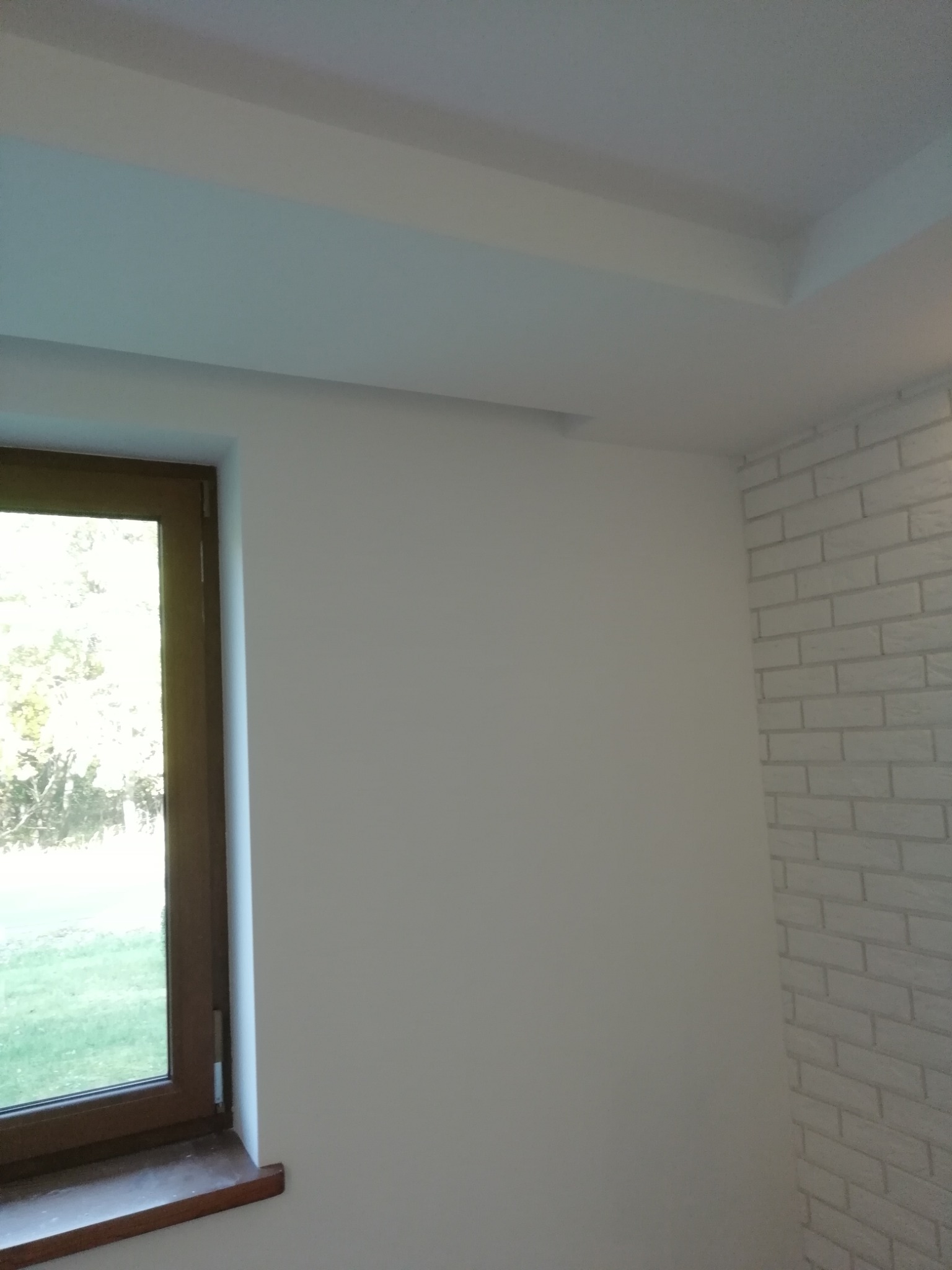 Świeżo pomalowane wnętrze z białymi ścianami, oknem z drewnianą ramą i fragmentem ściany z cegły pomalowanej na biało. Widoczny fragment sufitu z dekoracyjnym podwieszeniem.