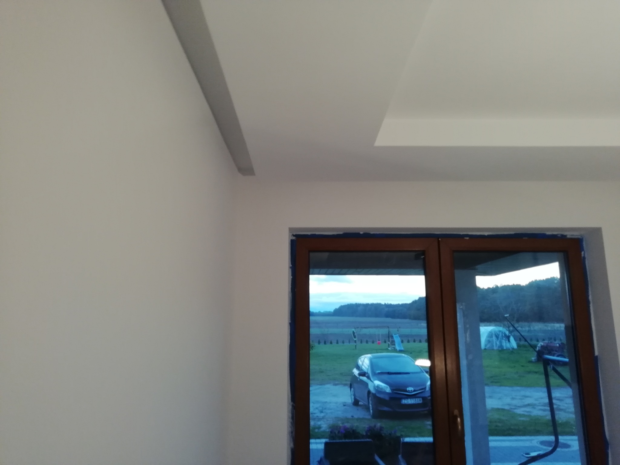 Świeżo pomalowane, białe ściany i sufit z widocznymi listwami dekoracyjnymi, okno z widokiem na podjazd z zaparkowanym samochodem i ogród.