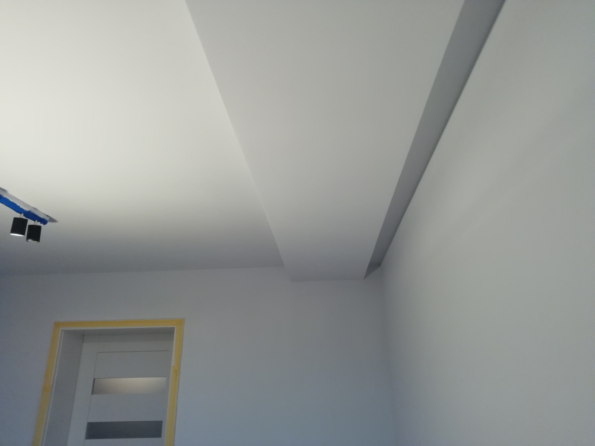 Pomalowane na biało wnętrze z fragmentem sufitu podwieszanego, widoczna listwa oświetleniowa i przygotowane do malowania ościeżnice drzwiowe z taśmą malarską.