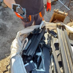 FHU Syl-Mar Sylwia Katarzyna Gabor - Pracownik w pomarańczowych rękawicach trzyma profil aluminiowy nad workiem pełnym aluminiowych odpadów, obok stoi mała koparka.