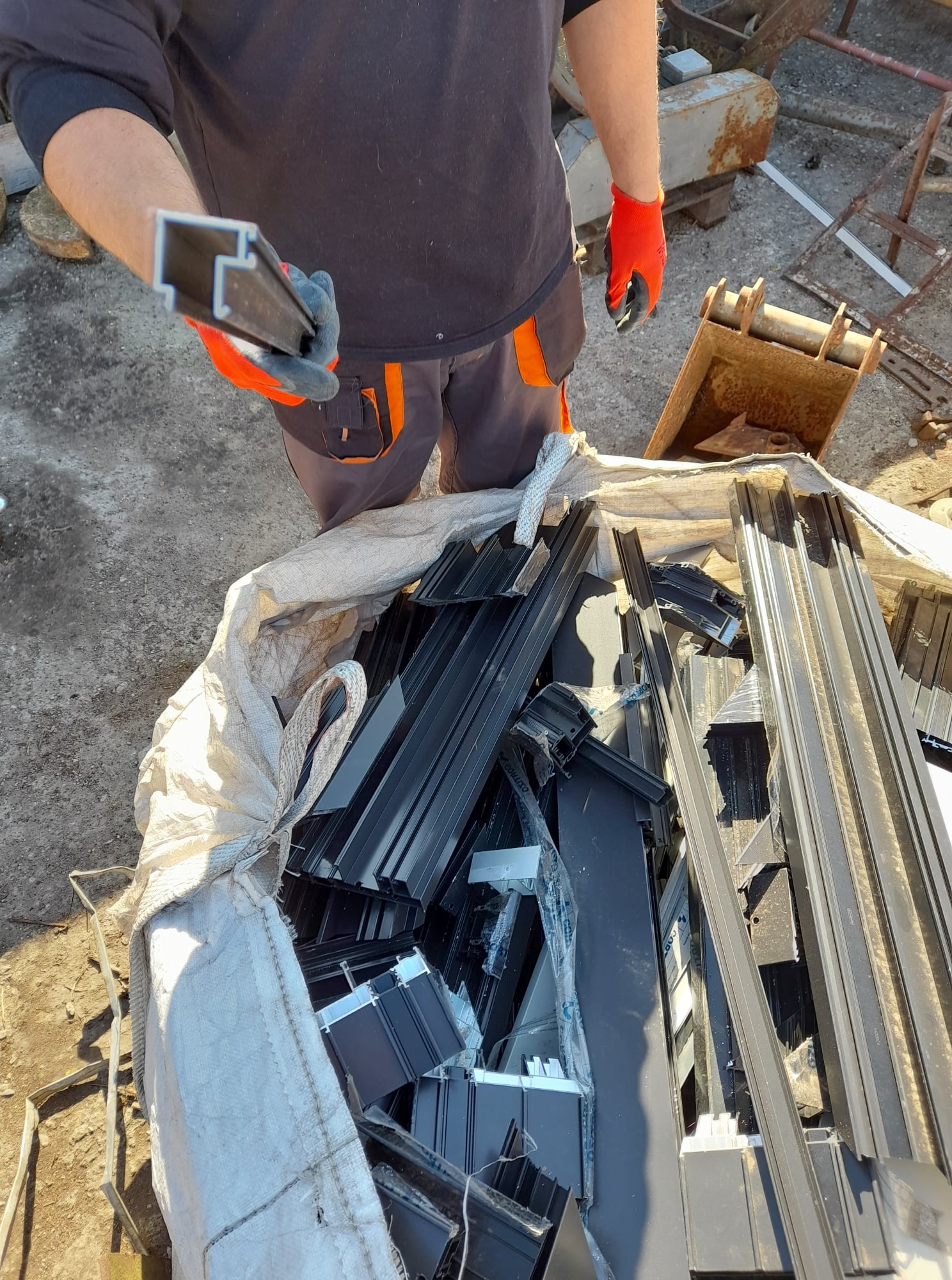 Pracownik w pomarańczowych rękawicach trzyma profil aluminiowy nad workiem pełnym aluminiowych odpadów, obok stoi mała koparka.