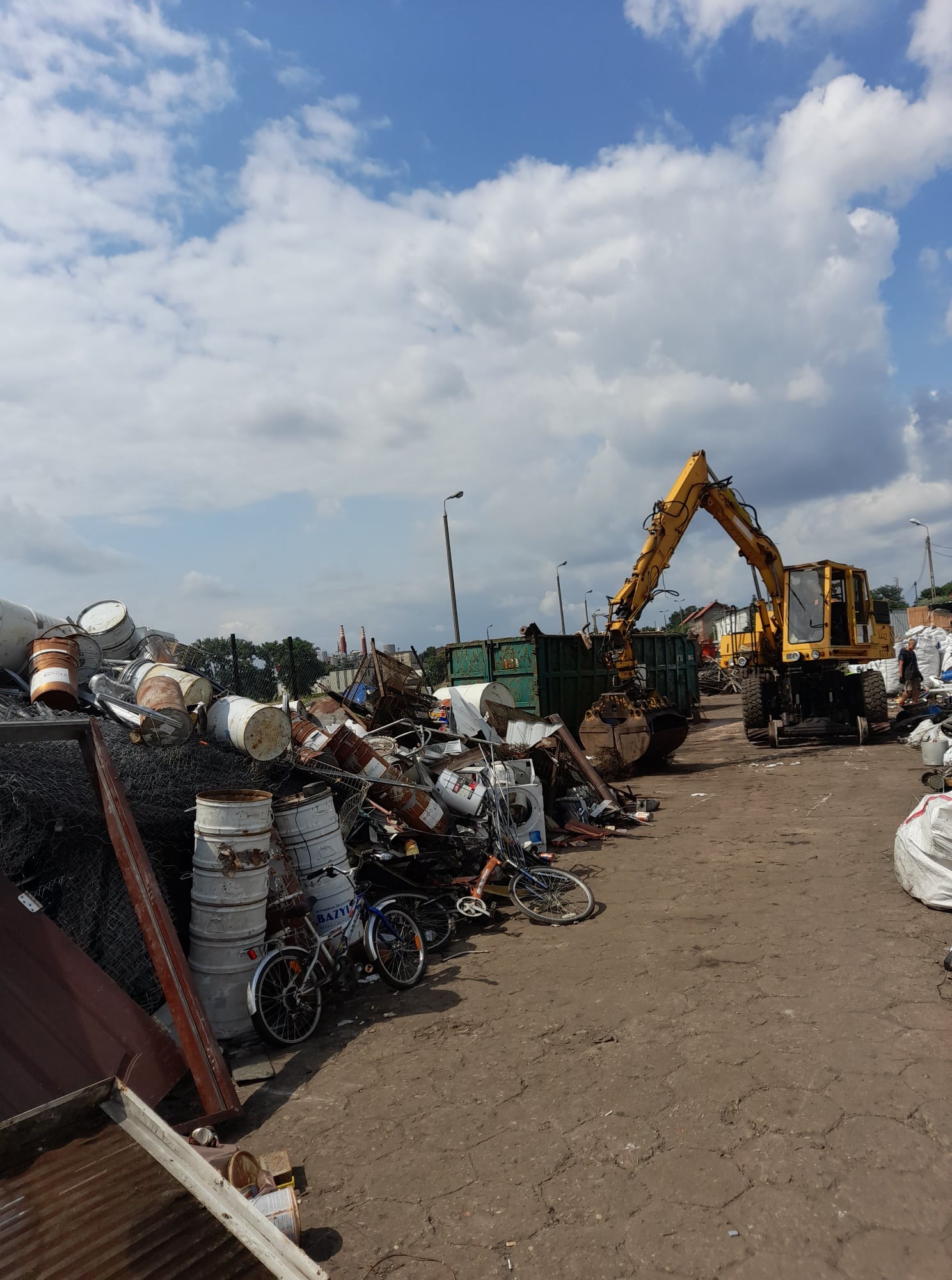 Żółta koparka załadowuje złom do kontenera, w tle sterta metalowych beczek, rowerów i innych odpadów na placu składowym pod błękitnym niebem.