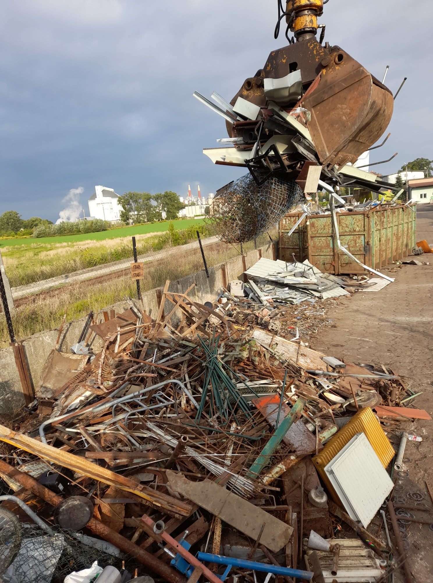 Żuraw z łyżką nabiera metalowe odpady, widoczny kontener, w tle krajobraz z budynkami przemysłowymi i pochmurnym niebem.