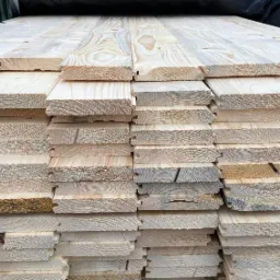 Stos jasnych desek z widocznymi słojami drewna i frezowanymi krawędziami, ułożonych równo jeden na drugim, z ciemnozielonym materiałem w tle.