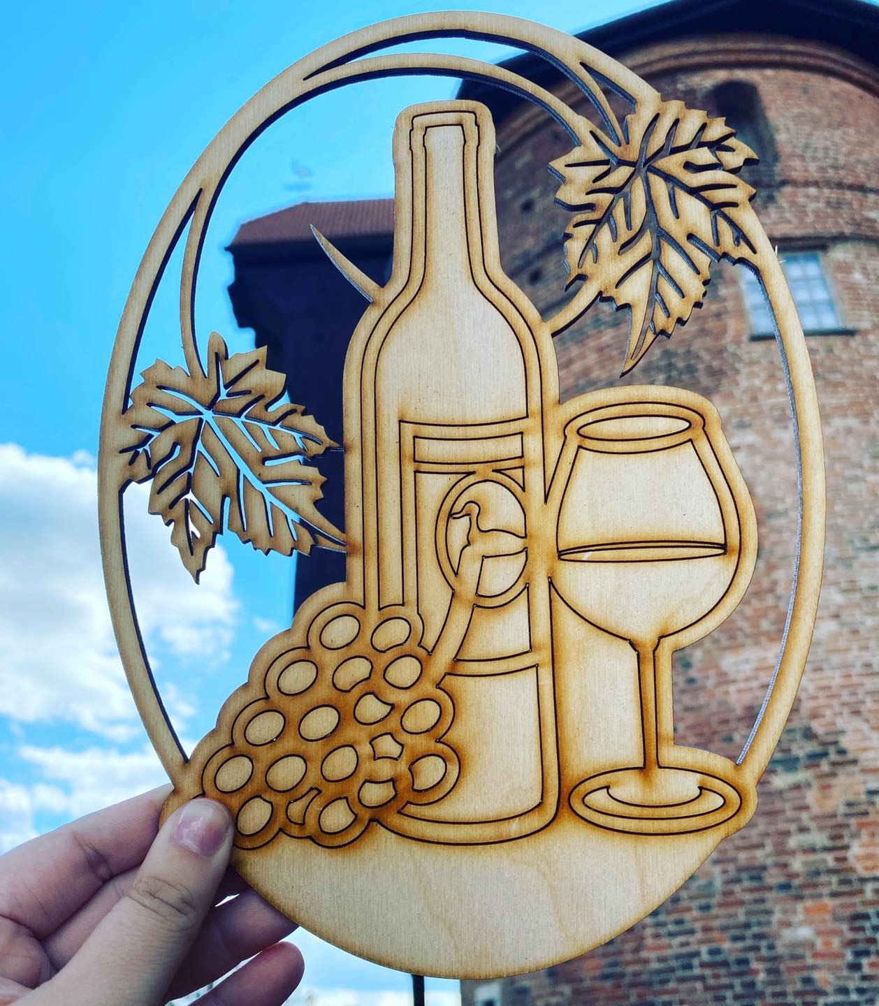 Drewniana dekoracja przedstawiająca butelkę wina, kieliszek i winogrona, trzymana na tle ceglanej wieży i błękitnego nieba.
