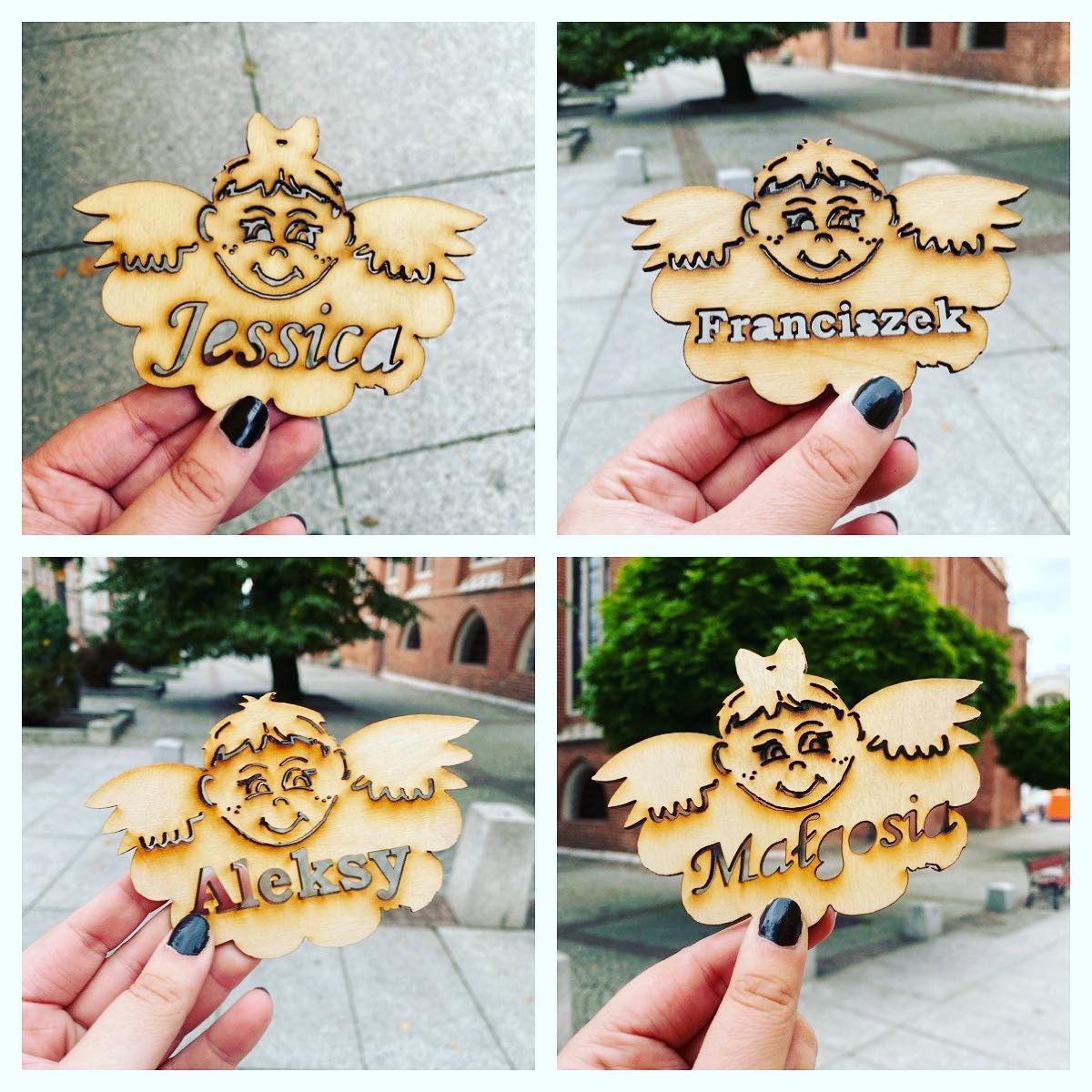 Cztery drewniane zawieszki w kształcie aniołów z wygrawerowanymi imionami Jessica, Franciszek, Aleksy i Małgosia, trzymane w dłoniach z ciemnym manicure na tle miejskiej architektury.