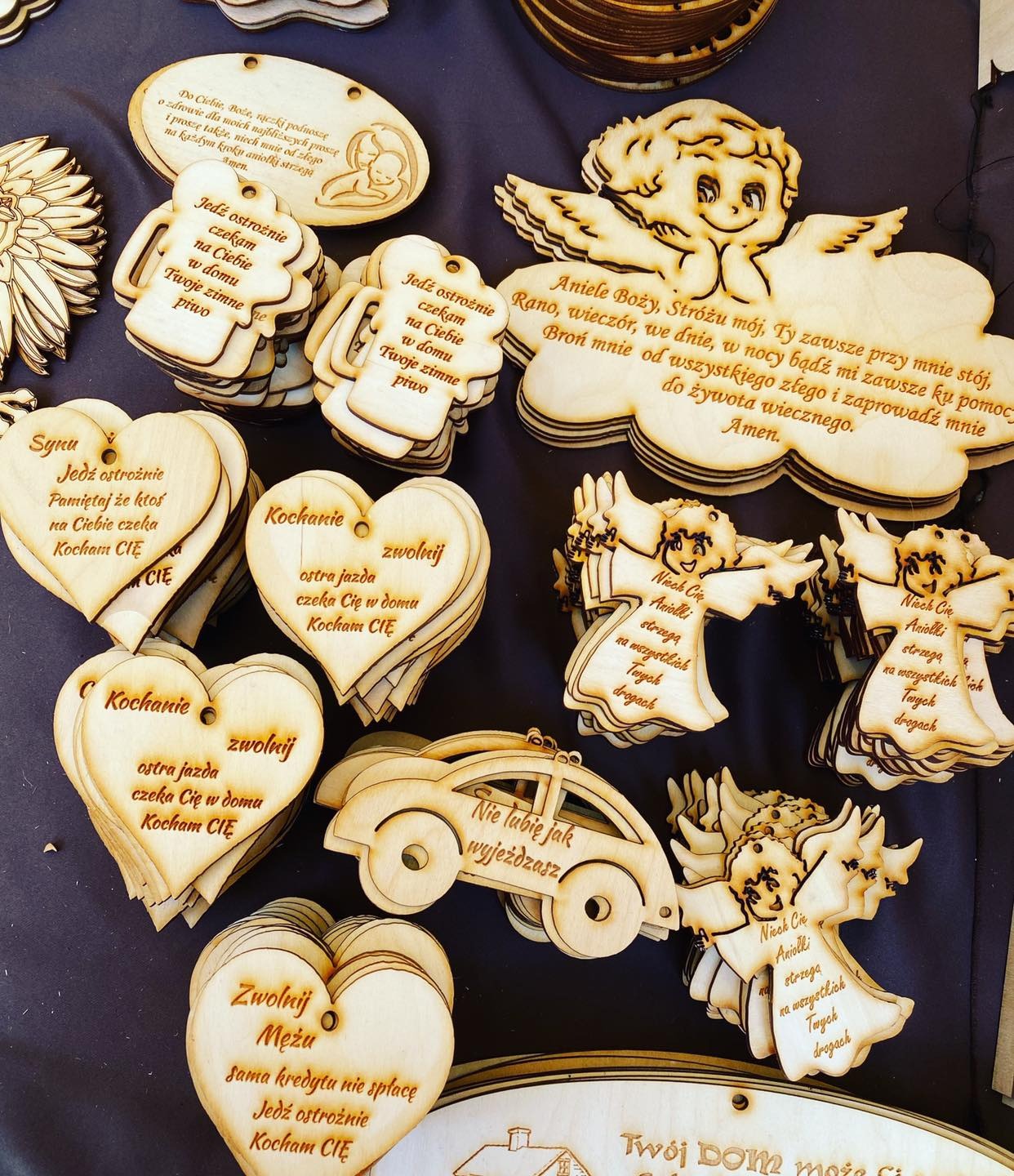 Drewniane ozdoby z grawerowanymi napisami, w tym serca, anioły i samochód, prezentowane na ciemnym tle w Karnicach.