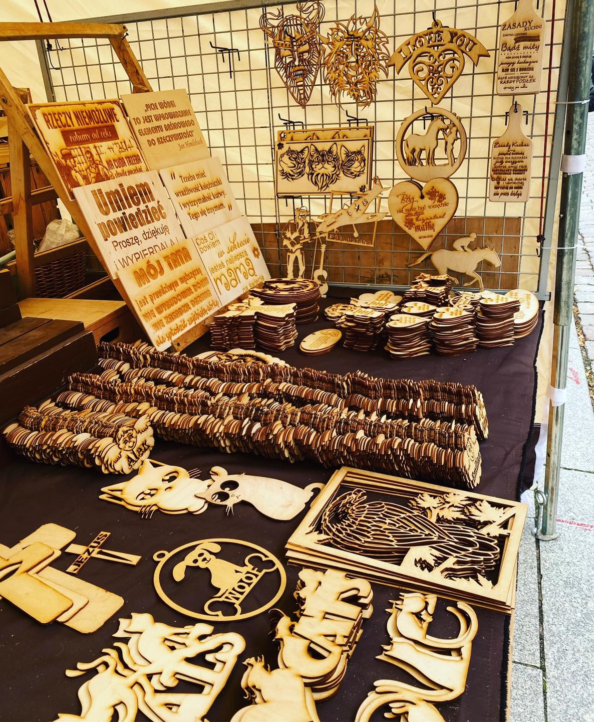Stoisko targowe z drewnianymi ozdobami i wycinankami, w tym zawieszki z motywami zwierząt, serca z napisami i tabliczki z cytatami, ekspozycja na brązowym obrusie i metalowej kratownicy.