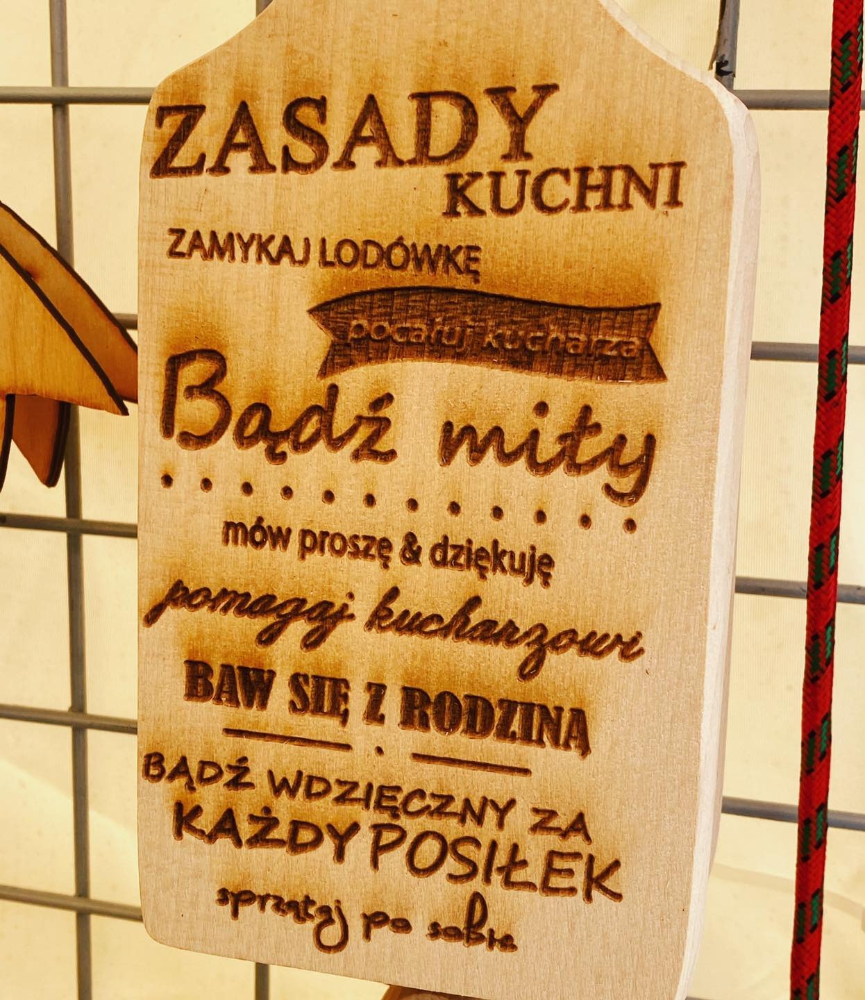 Drewniana tabliczka z wygrawerowanymi zasadami kuchni, wisząca na metalowej siatce, z widoczną czerwoną liną z boku.