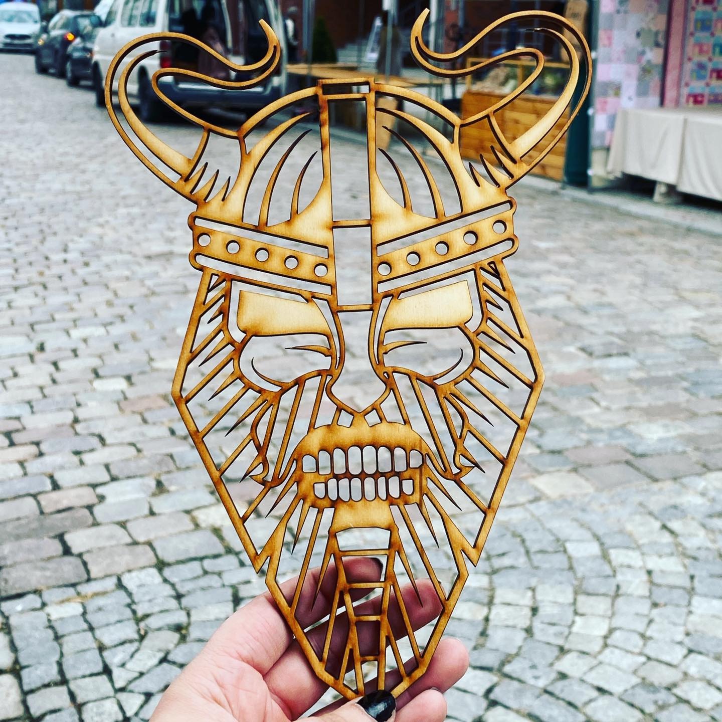 Drewniana, ażurowa maska wikinga z rogami, trzymana w dłoni na tle brukowanej ulicy.