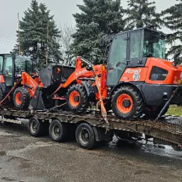 Transport trzech pomarańczowych ładowarek kołowych Kubota R090 na niskopodwoziowej platformie ciężarówki, zabezpieczonych pasami transportowymi, na tle pochmurnego nieba i zielonych drzew.