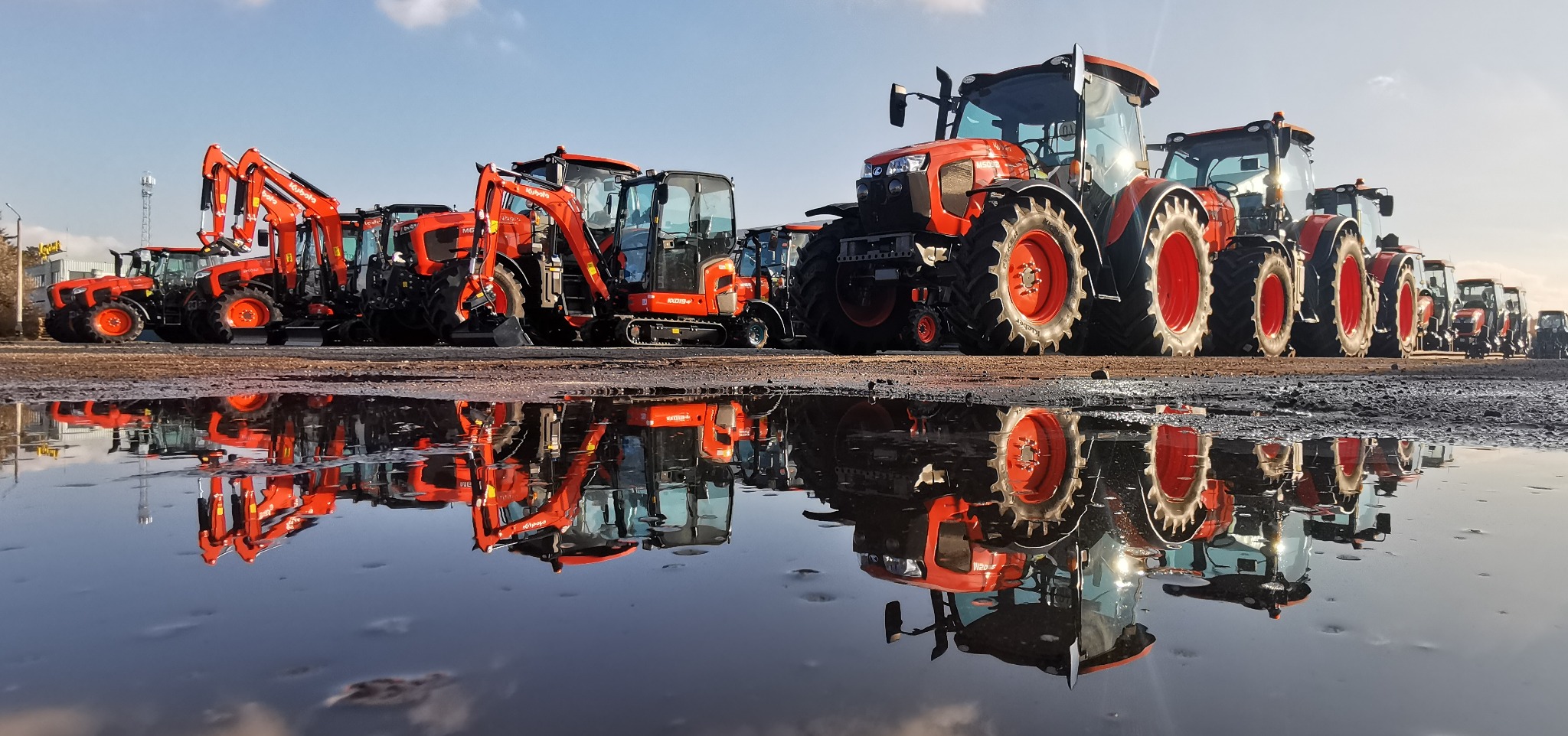 Rząd nowych, pomarańczowych maszyn budowlanych Kubota, w tym ciągniki i minikoparki, odbijających się w dużej kałuży na ziemi.