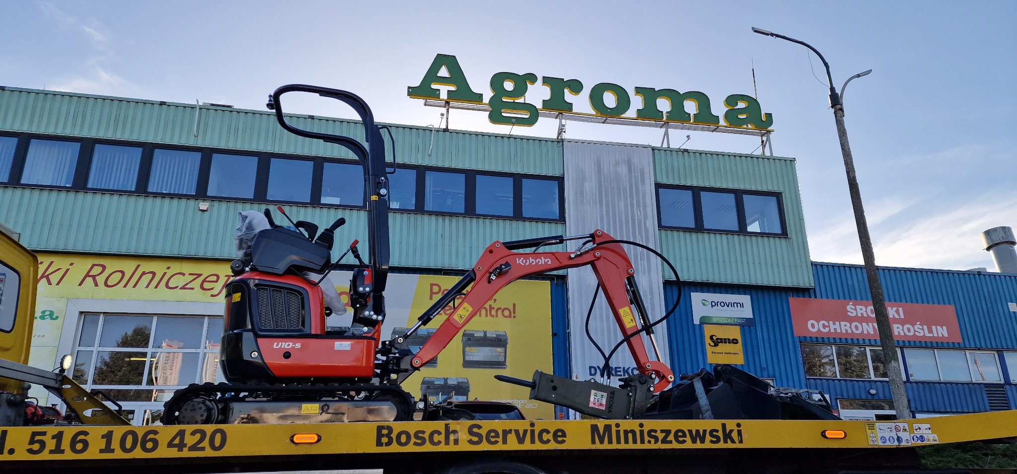 Czerwona minikoparka Kubota U10-5 na lawecie Bosch Service Miniszewski w Warszawie, na tle budynku z napisem Agroma.