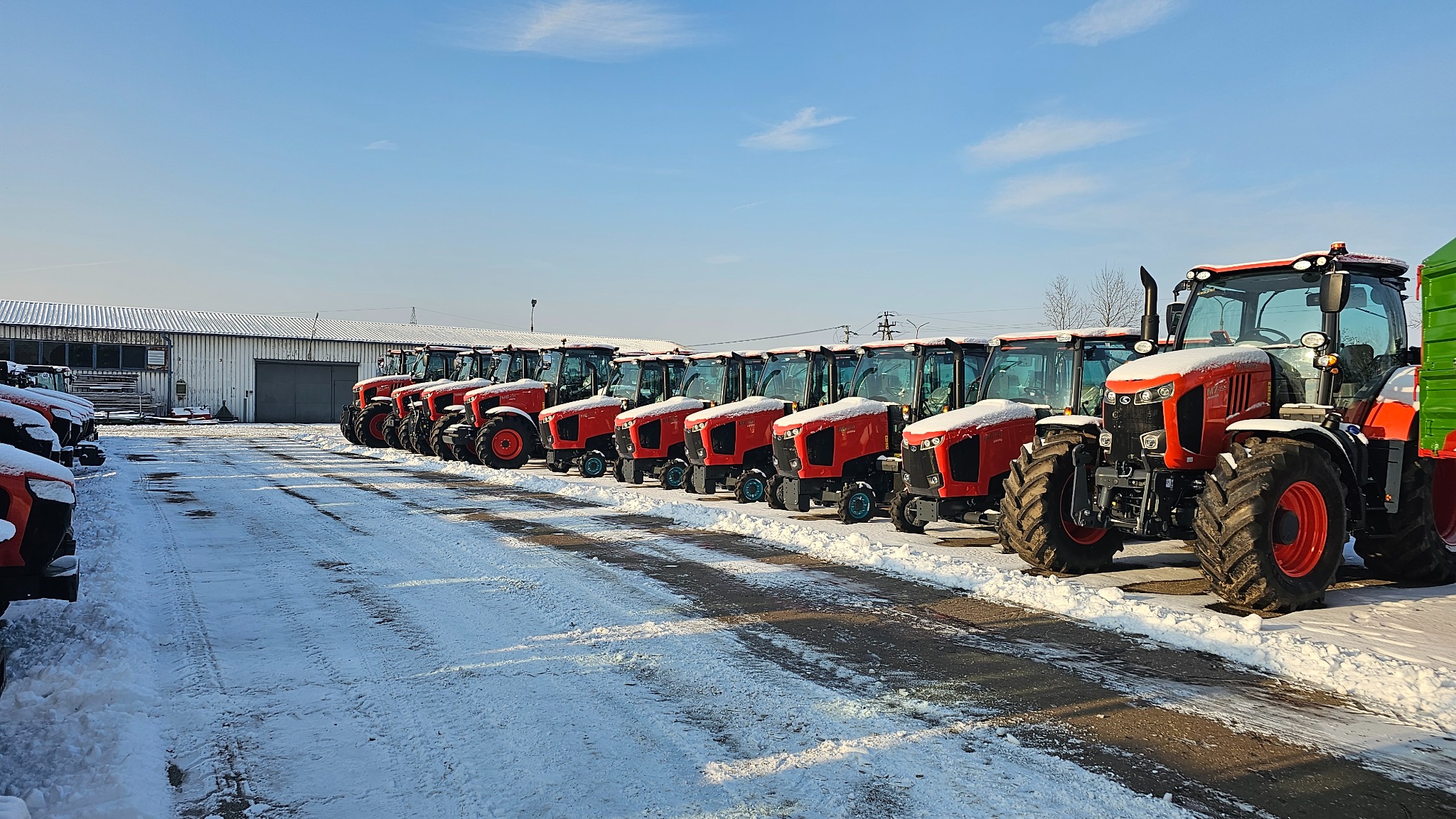 Rząd nowych, czerwonych traktorów Kubota M7152 na śniegu przed magazynem, w zimowym słońcu.
