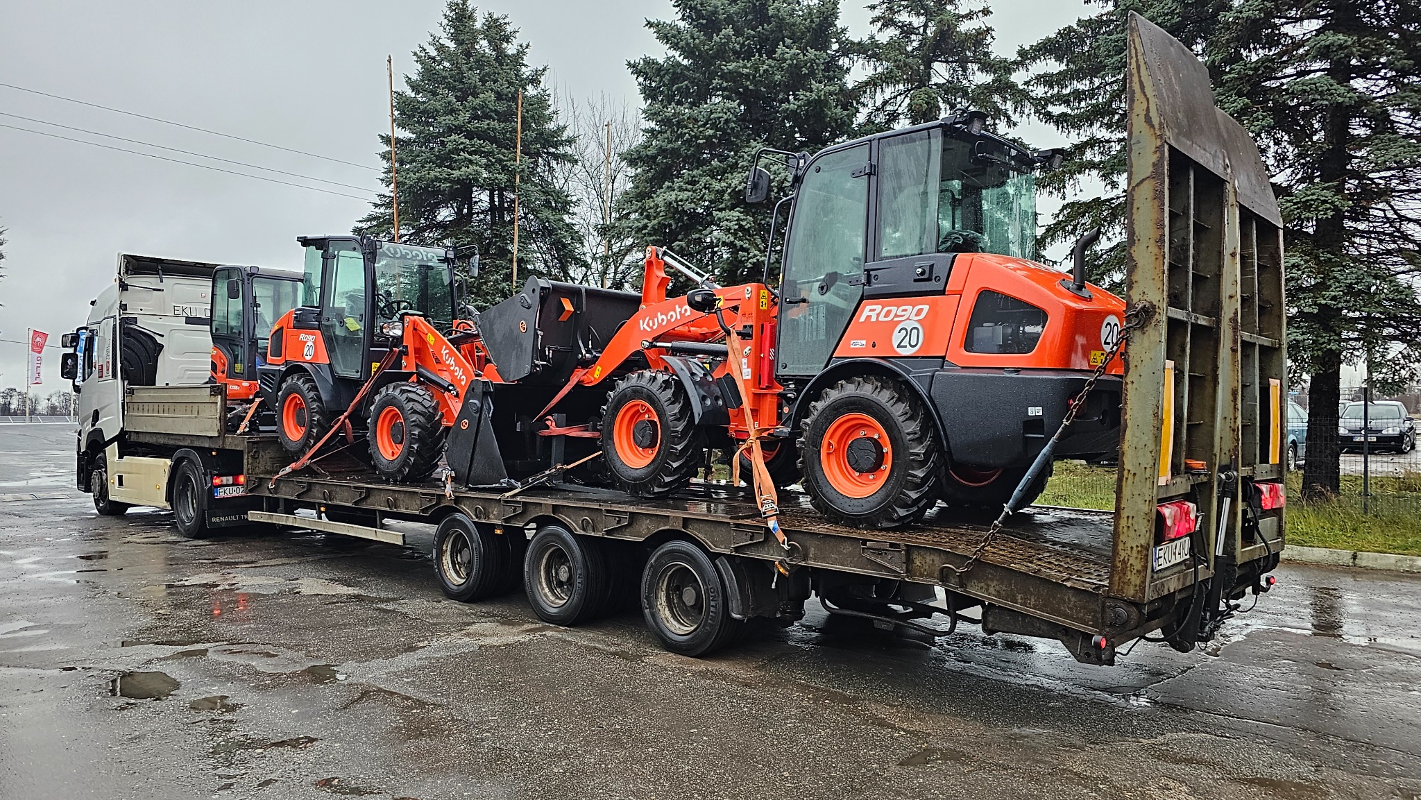 Transport trzech pomarańczowych ładowarek kołowych Kubota R090 na niskopodwoziowej platformie ciężarówki, zabezpieczonych pasami transportowymi, na tle pochmurnego nieba i zielonych drzew.