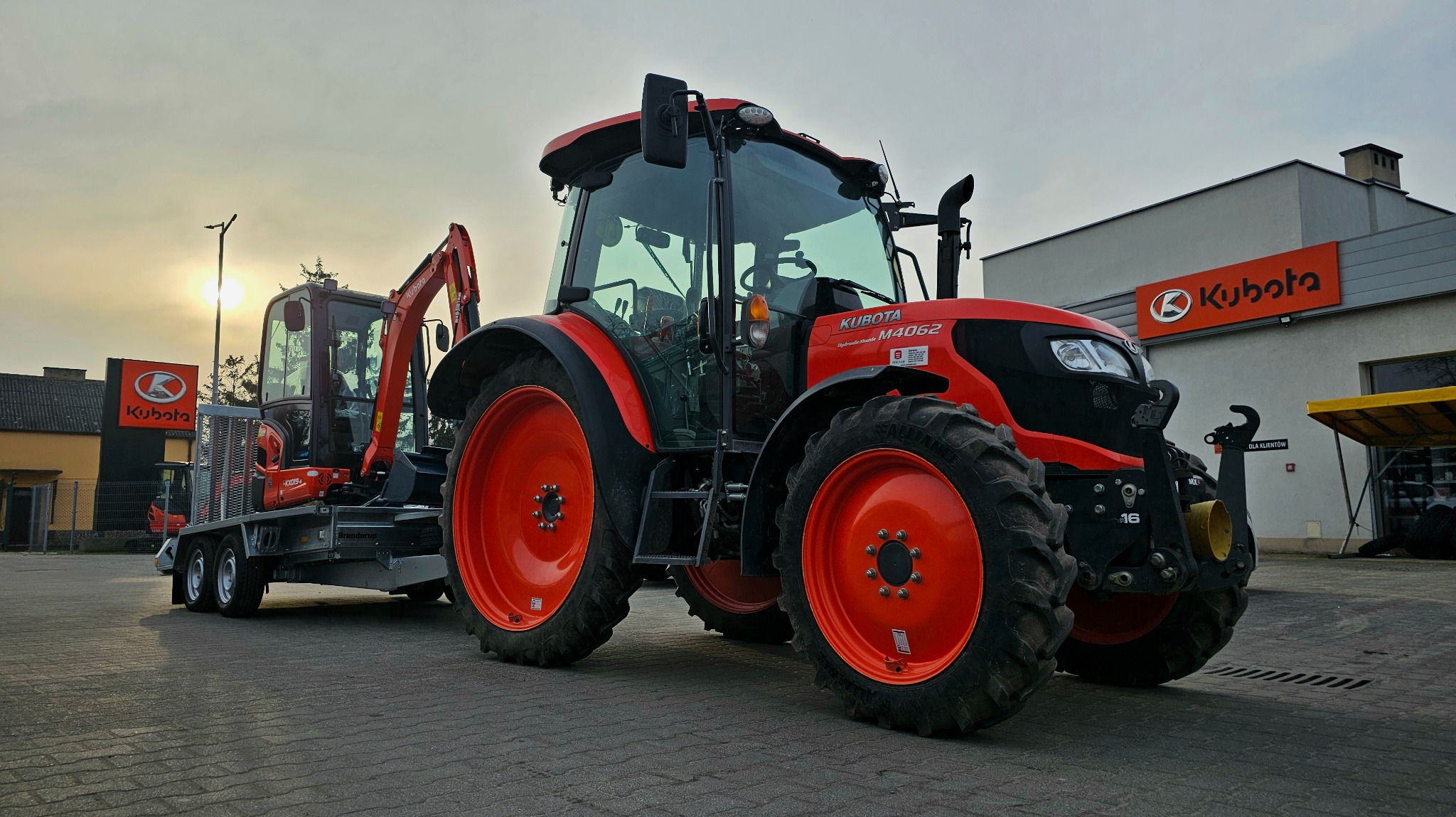 Czerwony ciągnik Kubota M4062 z przyczepą, na której stoi minikoparka Kubota, na tle budynku z logo Kubota.
