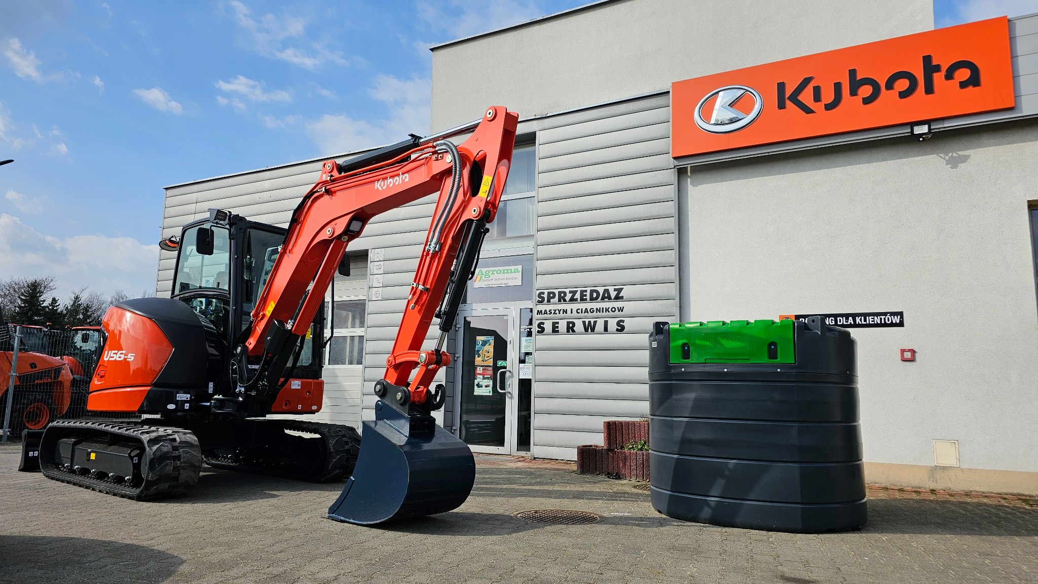 Pomarańczowa minikoparka Kubota U56-5 przed budynkiem firmy Agroma z logo Kubota na dachu, obok zbiornik na paliwo.