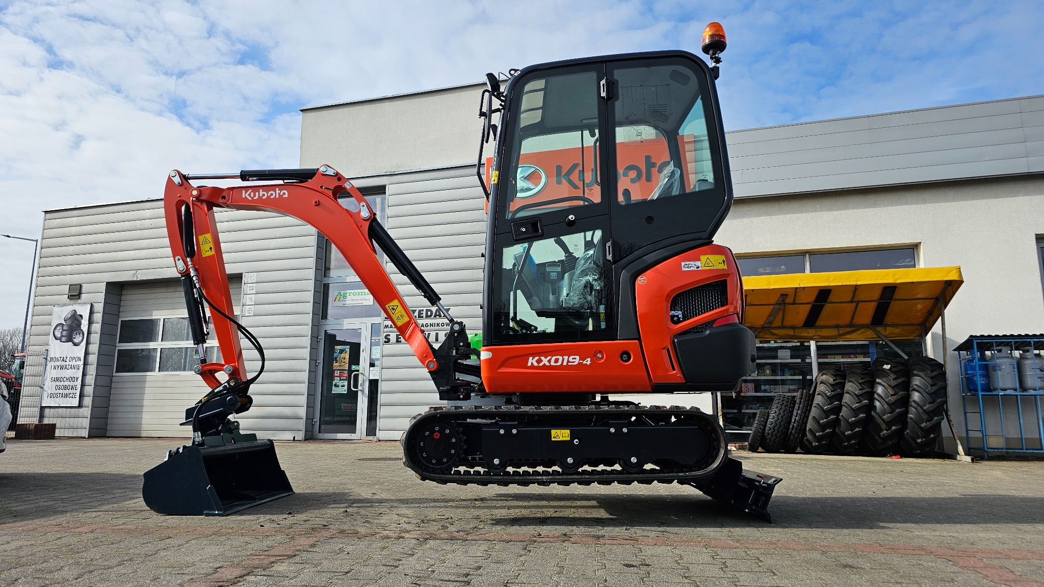 Pomarańczowa minikoparka Kubota KX019-4 z czarną kabiną i łyżką, prezentowana na placu przed budynkiem z oknami i żółtym zadaszeniem, widoczne opony i butle gazowe.