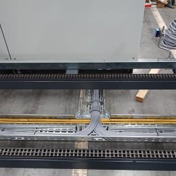 S-Automatic Sławomir Słomiński - Szyna kablowa z ułożonymi przewodami w szarych peszlach, widok z góry, fragment szafy sterowniczej w tle, zwój kabli na podłodze obok.