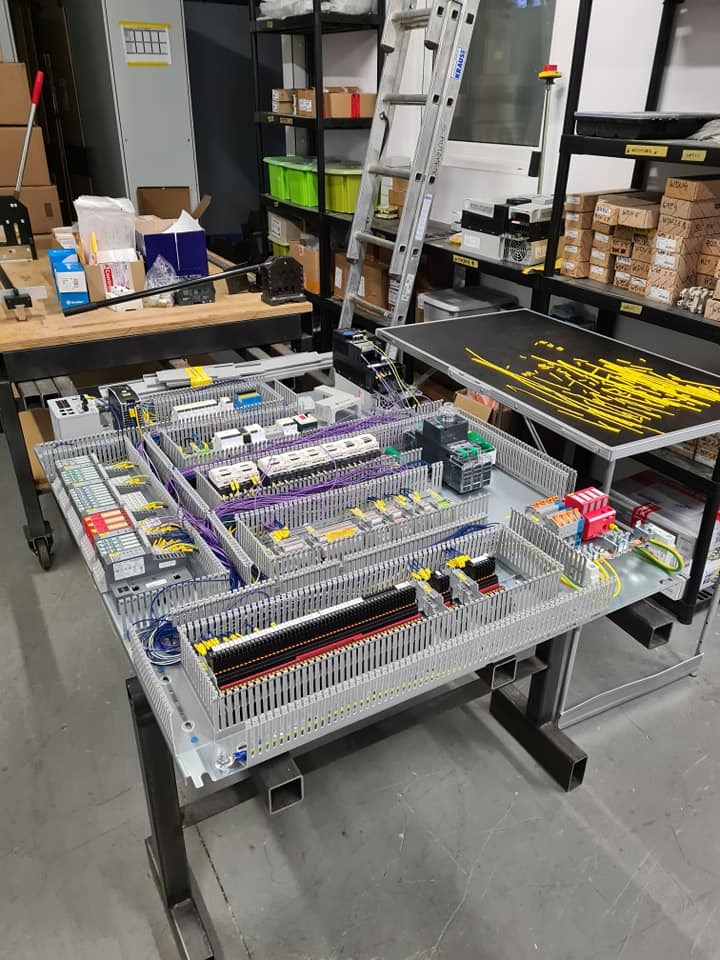 Rozbudowany panel sterowania automatyki z licznymi przekaźnikami, przewodami i elementami elektrycznymi, umieszczony na metalowym stole w warsztacie, z drabiną i regałami w tle.