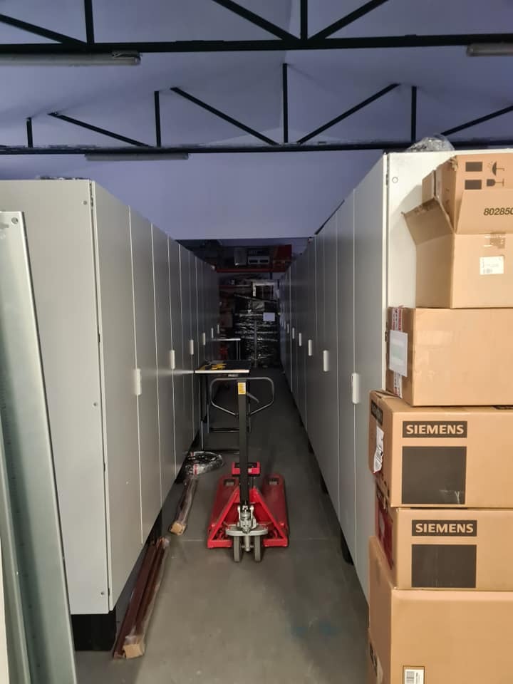 Wózek paletowy w wąskim korytarzu między regałami magazynowymi z kartonami Siemens na jednym z nich.