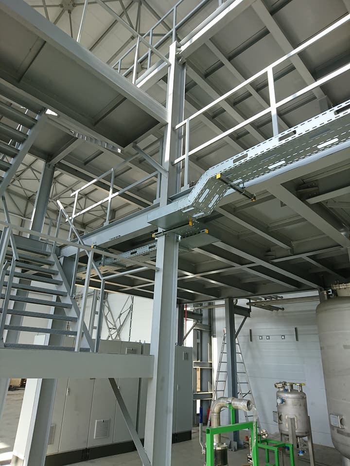 Wnętrze hali przemysłowej z metalową konstrukcją nośną, schodami, korytkami kablowymi i zbiornikami ciśnieniowymi, oświetlone naturalnym światłem.