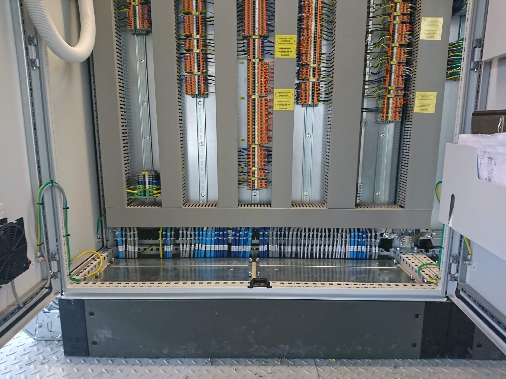 Otwarta szafa sterownicza z widocznymi licznymi przewodami, listwami zaciskowymi i modułami elektronicznymi, wentylator po lewej stronie, podłoga metalowa z fakturą.