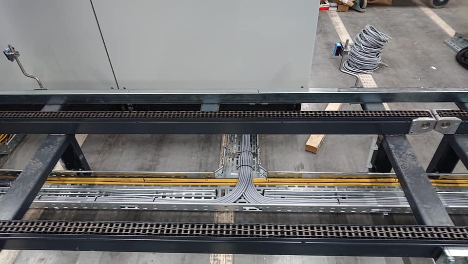 Szyna kablowa z ułożonymi przewodami w szarych peszlach, widok z góry, fragment szafy sterowniczej w tle, zwój kabli na podłodze obok.