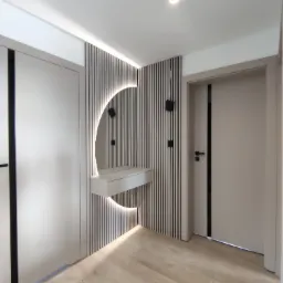 Nowoczesny przedpokój z zabudową: szafa z czarnymi wstawkami, ściana z pionowych lameli z podświetlanym lustrem w kształcie półksiężyca i konsolą, drewniana podłoga, oświetlenie punktowe.