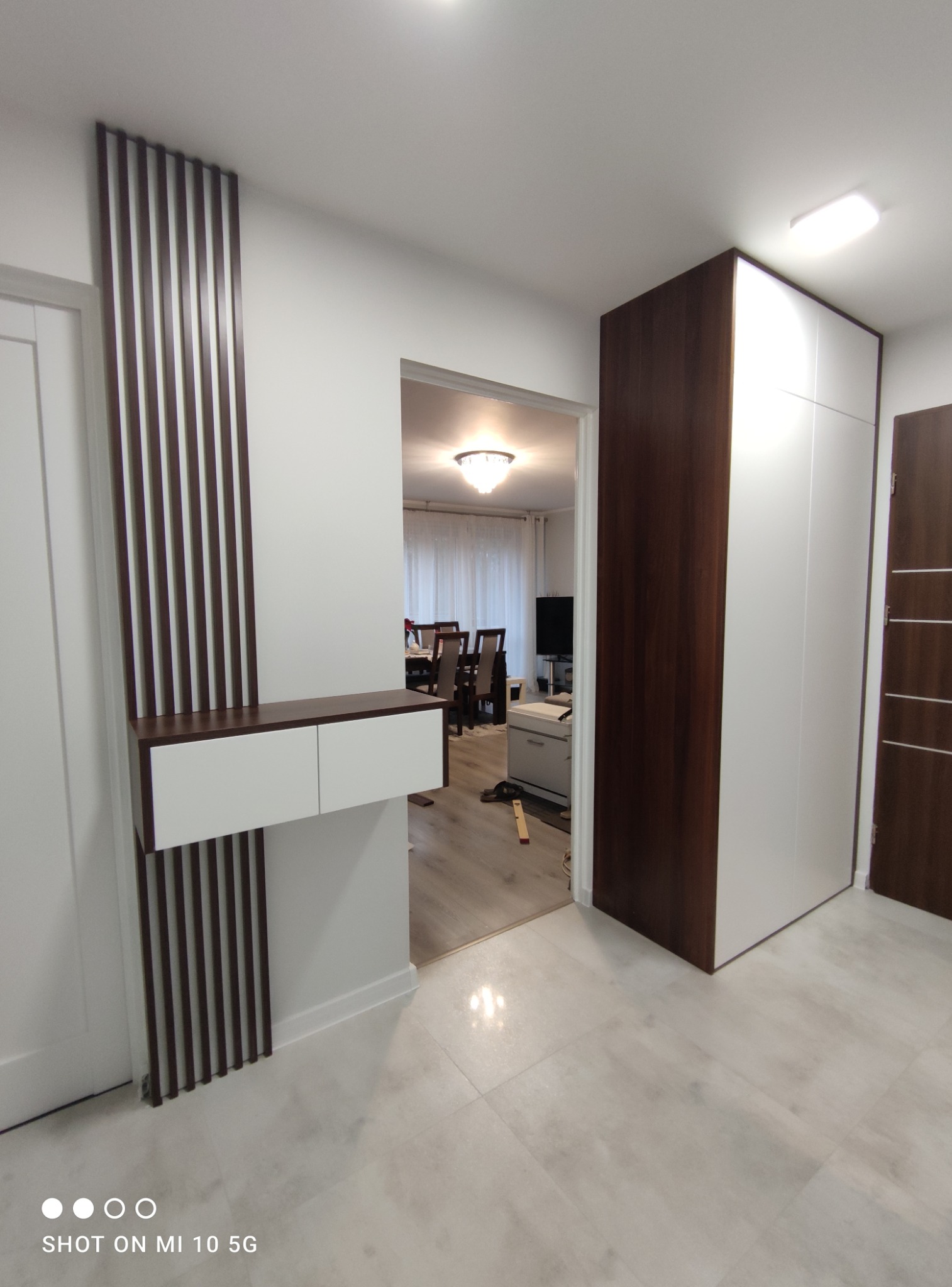 Nowoczesna aranżacja przedpokoju w Trzebini z zabudową szafy wnękowej w kolorze białym i ciemnego drewna oraz dekoracyjnym panelem ściennym z listewek i wiszącą szafką z szufladami.