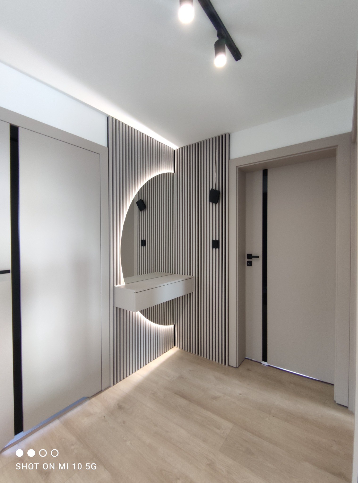 Nowoczesny przedpokój z zabudową: szafa z czarnymi wstawkami, ściana z pionowych lameli z podświetlanym lustrem w kształcie półksiężyca i konsolą, drewniana podłoga, oświetlenie punktowe.