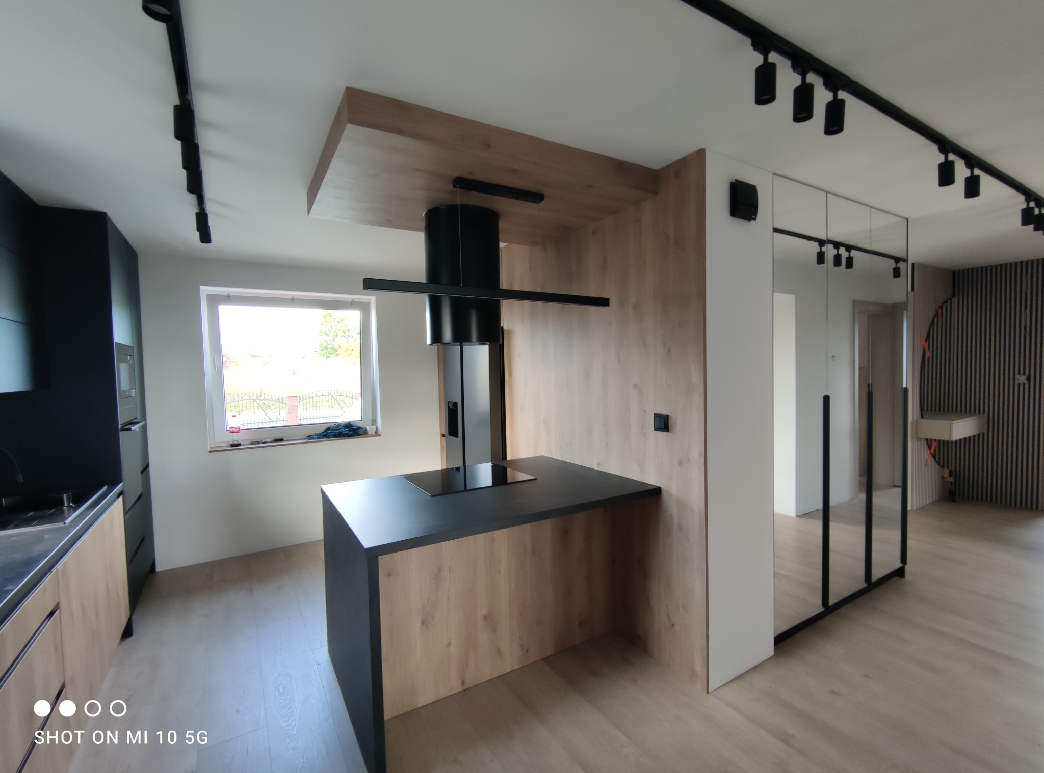 Nowoczesna kuchnia z wyspą i okapem cylindrycznym, wykończona fornirem drewnopodobnym, oświetlenie szynowe sufitowe, minimalistyczny design.