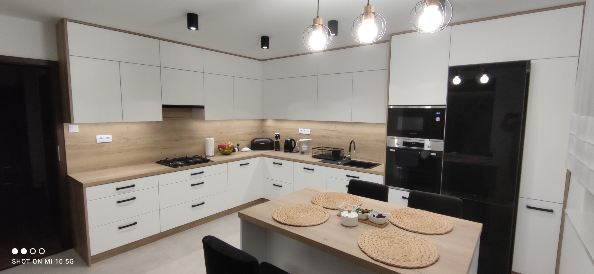 Jasna kuchnia w kształcie litery L z białymi frontami i drewnianym blatem, oświetlona wiszącymi lampami z żarówkami Edison, widoczny stół z podkładkami i przekąskami, czarny sprzęt AGD w zabudowie.