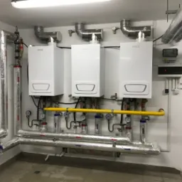 Trzy białe, wiszące piece grzewcze z podłączeniami rur w izolacji aluminiowej i żółtej rury gazowej, zainstalowane na białej ścianie w pomieszczeniu technicznym.