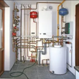 Złożona instalacja centralnego ogrzewania z piecem, zasobnikiem ciepłej wody i licznymi rurami miedzianymi w pomieszczeniu z oknem i regałem.