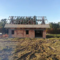 Budynek w trakcie budowy z widoczną konstrukcją dachu z drewnianych krokwi, murowane ściany z czerwonej cegły, teren wokół z widocznymi śladami opon i drzewami w tle.