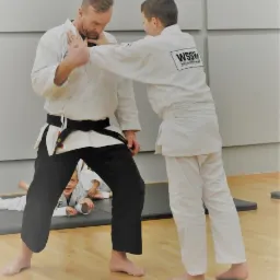 Instruktor w białym kimonie z czarnym pasem demonstruje technikę obrony na chłopcu w białym kimonie, w tle ćwiczące dzieci.