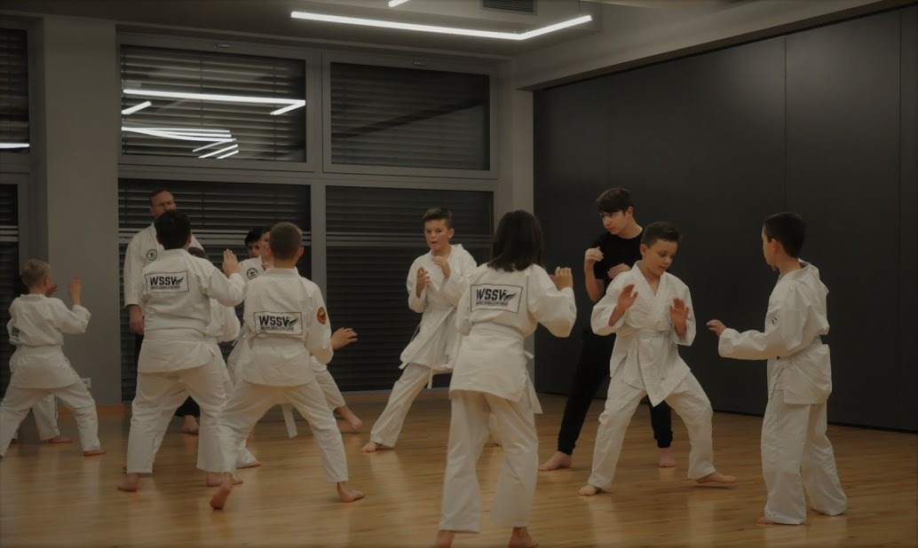 Grupa dzieci w białych kimonach ćwiczy karate na drewnianej podłodze w jasnym pomieszczeniu z dużymi oknami i trenerem w tle.