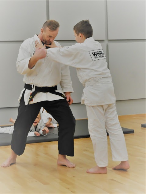 Instruktor w białym kimonie z czarnym pasem demonstruje technikę obrony na chłopcu w białym kimonie, w tle ćwiczące dzieci.