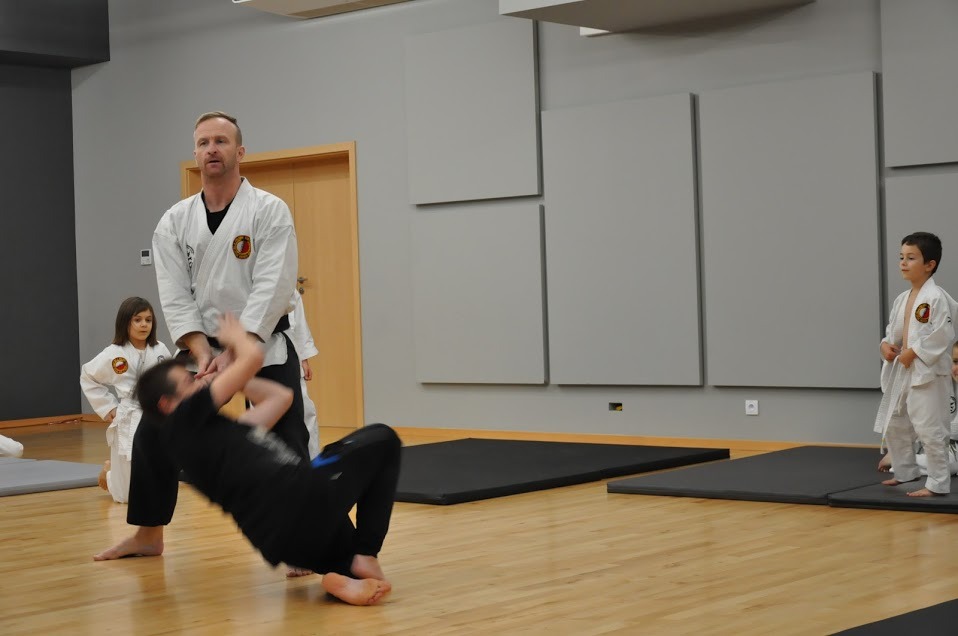 Instruktor w białym kimonie demonstruje technikę upadku na macie podczas zajęć z dziećmi w sali treningowej.