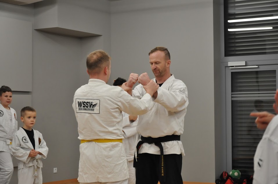Instruktor w białym kimonie z czarnym pasem demonstruje technikę walki wręcz innemu mężczyźnie w kimonie, na tle grupy uczniów w sali treningowej.