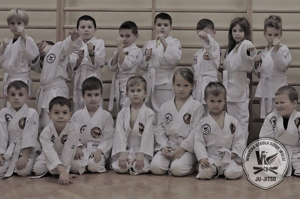 Grupa dzieci w białych kimonach pozuje do zdjęcia w sali treningowej, widoczne logo szkoły sztuk walki w prawym dolnym rogu.