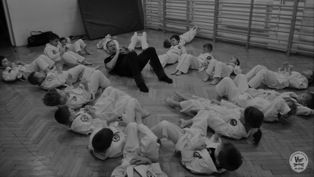 Grupa dzieci w białych kimonach leżących na podłodze wokół mężczyzny w czarnym ubraniu, który siedzi i patrzy w telefon, podczas zajęć z jiu-jitsu w sali gimnastycznej.