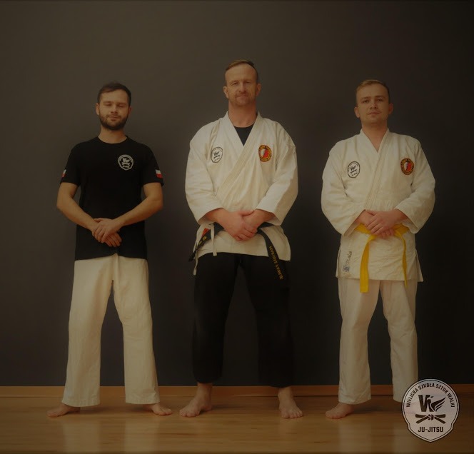 Trzech mężczyzn w strojach do sztuk walki: jeden w czarnej koszulce z logo, drugi w białym kimonie z czarnym pasem i trzeci w białym kimonie z żółtym pasem, stojący boso na drewnianej podłodze...