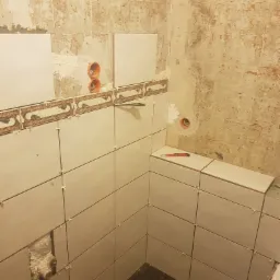 Ściany łazienki w trakcie układania jasnych płytek ceramicznych z dekoracyjnym pasem. Widoczne krzyżyki dystansowe, puszki elektryczne i nóż do cięcia płytek.