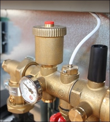 Mosiężny zawór odpowietrzający z czerwonym pokrętłem, manometrem i białym przewodem na tle metalowej ściany.