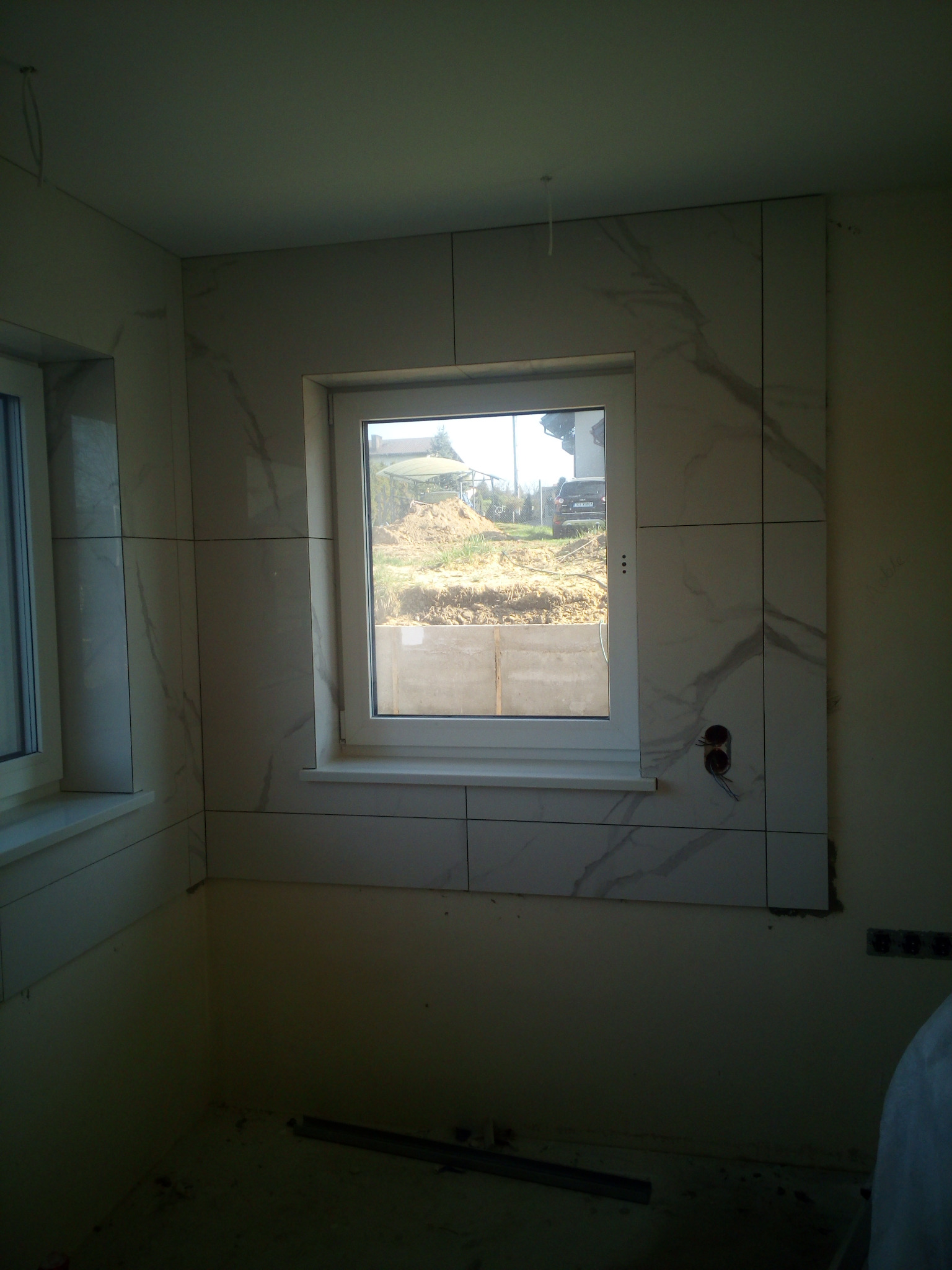 Wnętrze w trakcie wykańczania, widoczne białe płytki imitujące marmur na ścianach wokół okna, niedokończone gniazdka elektryczne i surowy stan podłogi.