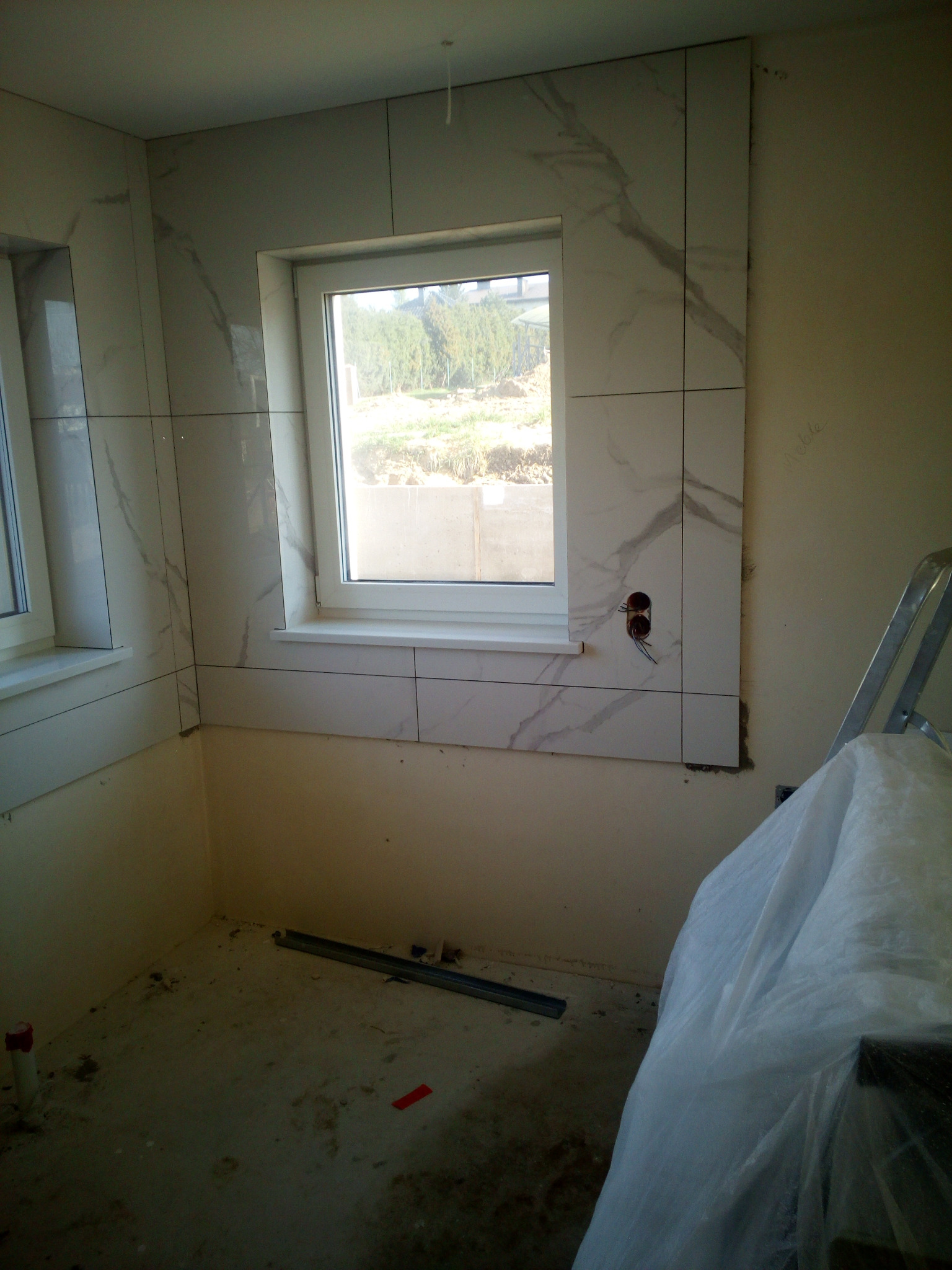 Fragment łazienki w trakcie prac wykończeniowych z oknem, ściany częściowo wyłożone białymi płytkami imitującymi marmur, widoczne puszki elektryczne i drabina zabezpieczona folią malarską.